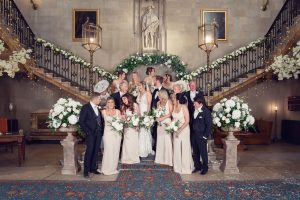 A large wedding party poses on a grand staircase adorned with white flowers and greenery at one of the city's historic wedding venues. The bride and groom, surrounded by their formally-clad friends, are beautifully captured by a fine art wedding photographer.