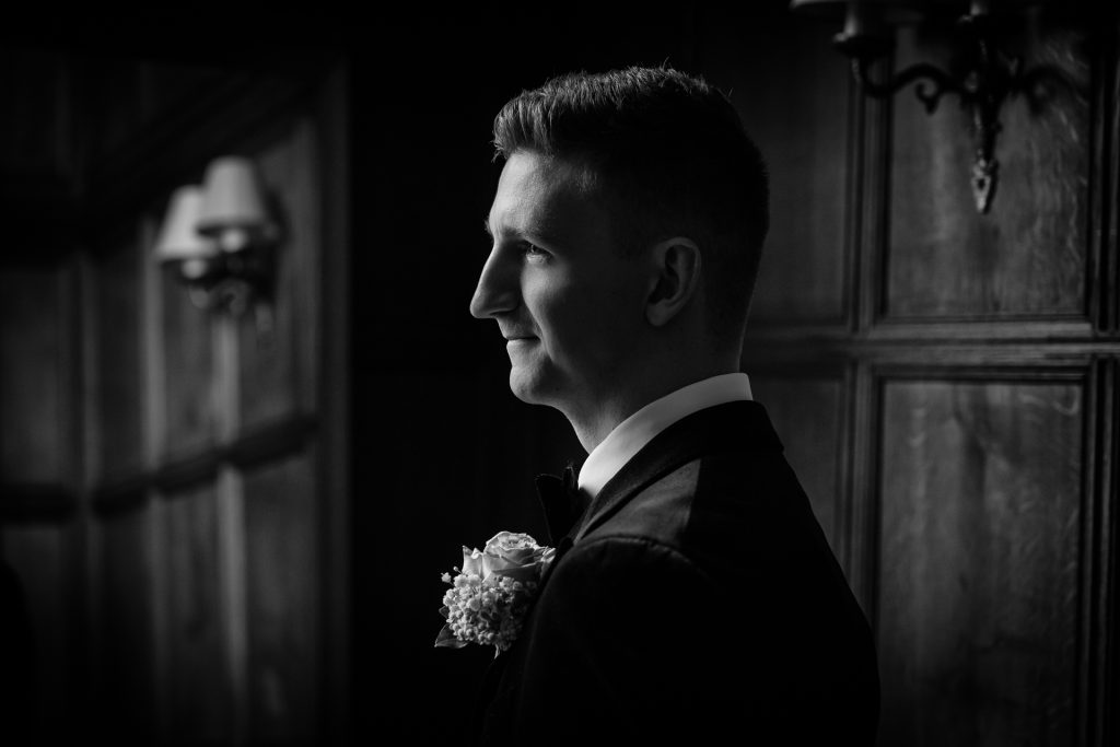 A man in a suit with a boutonniere stands in profile, smiling softly. The black and white photo has dramatic lighting and a classic wood-paneled background with a wall sconce visible.