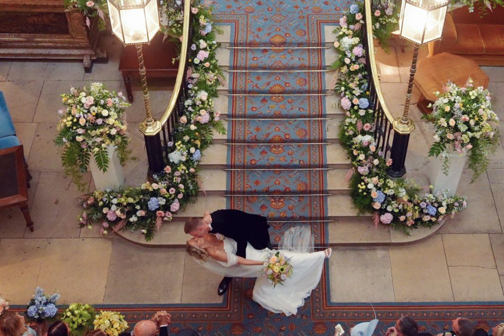 A bride and groom share a kiss at the base of ornate, flower-lined stairs in a grand venue, viewed from above. The bride holds a bouquet and dips back as the groom embraces her. Guests are seated nearby.