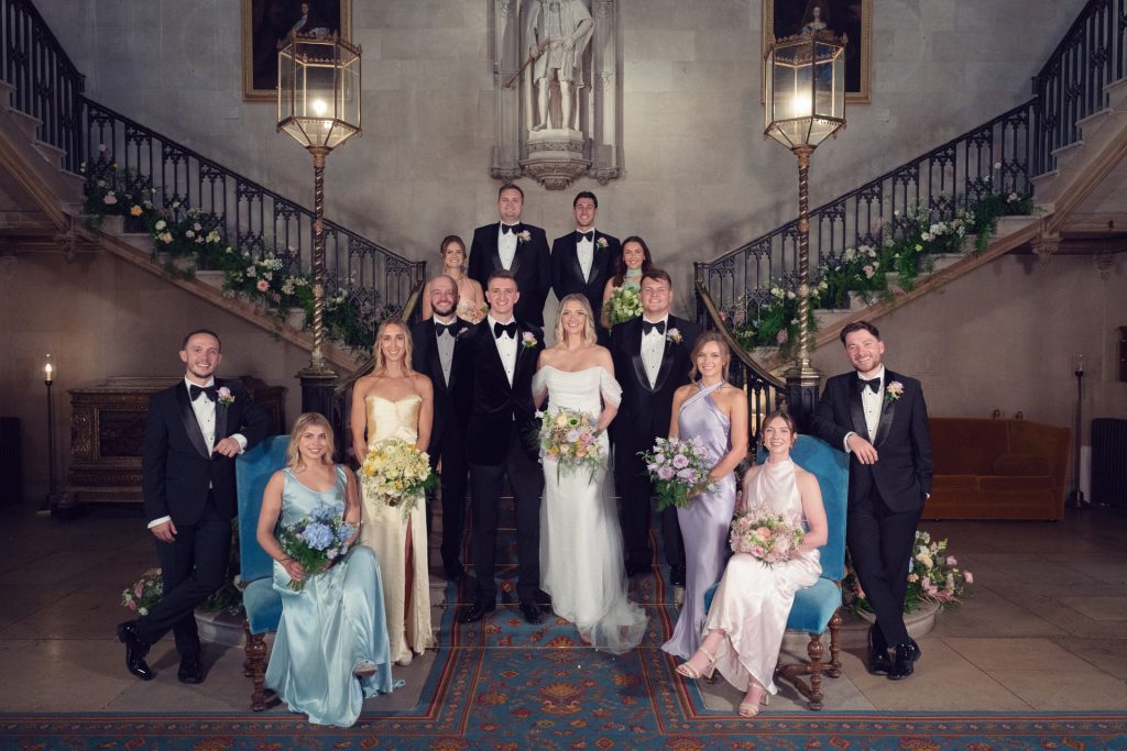 A wedding party poses indoors on a grand staircase. The bride, groom, bridesmaids, and groomsmen wear formal attire, holding bouquets and smiling. The setting features flowers, large lanterns, and an ornate carpet.
