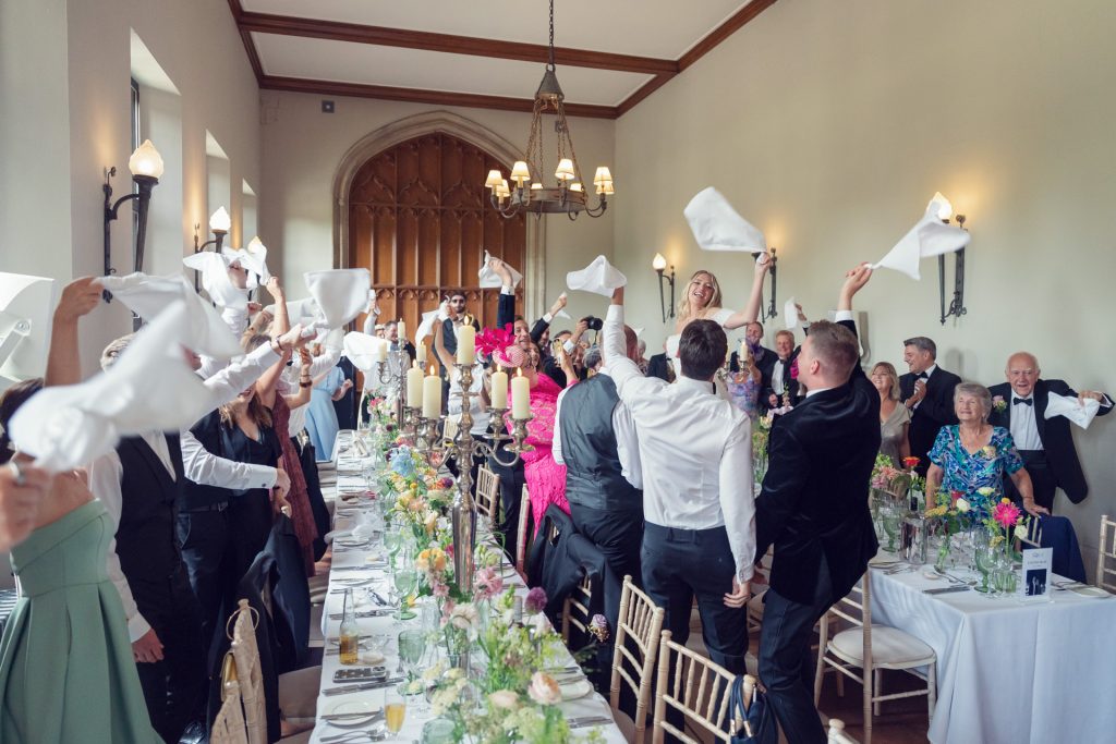 Wedding guests stand around a long, flower-decorated table, waving white napkins in celebration inside a bright, elegant hall with high ceilings and large arched doors.