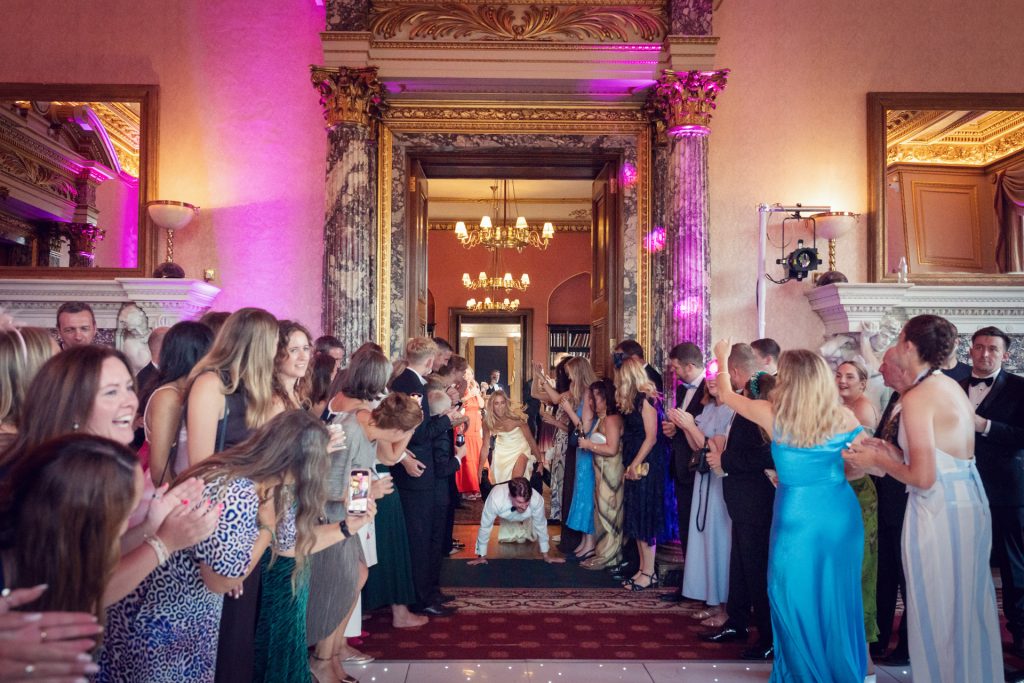A group of people in formal attire form two lines in a decorated hall, clapping and cheering as a person in white crawls on the floor between them toward the camera. Chandeliers and ornate columns are visible in the background.