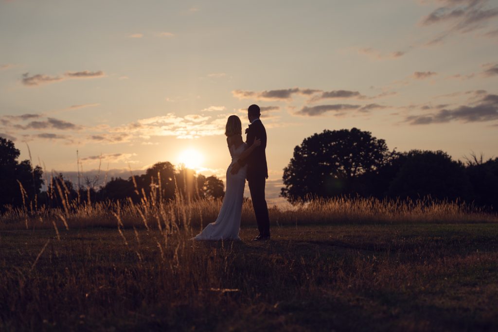 A bride and groom stand together in a grassy field at sunset, silhouetted against a golden sky with scattered clouds and distant trees.
