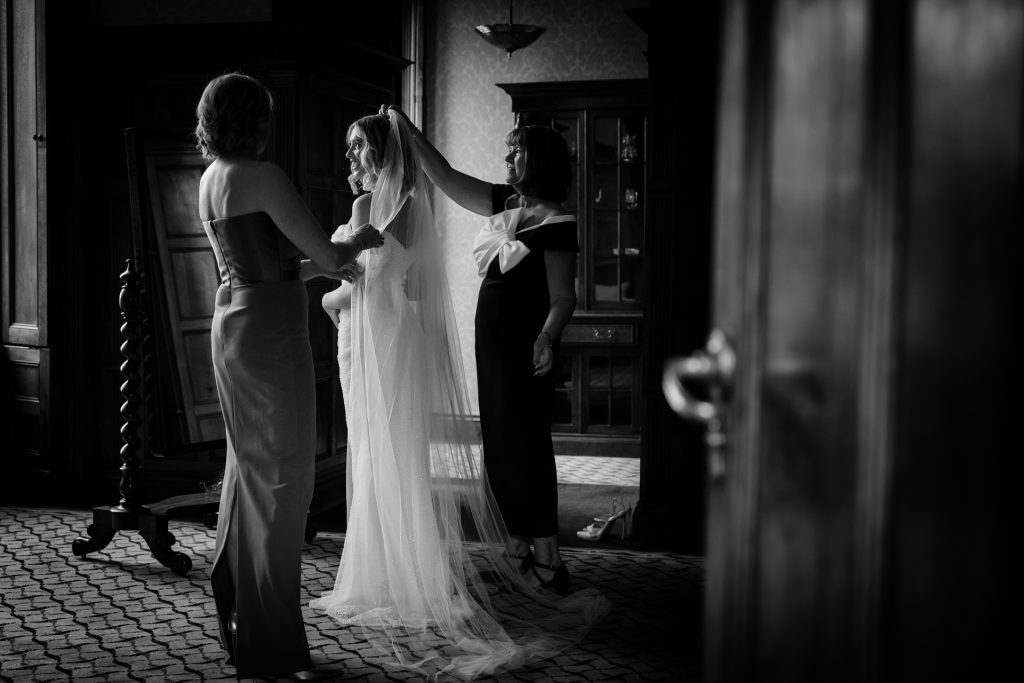 A bride in a veil stands inside a dimly lit room as two women help adjust her dress. The scene captures an intimate, candid moment before a wedding.
