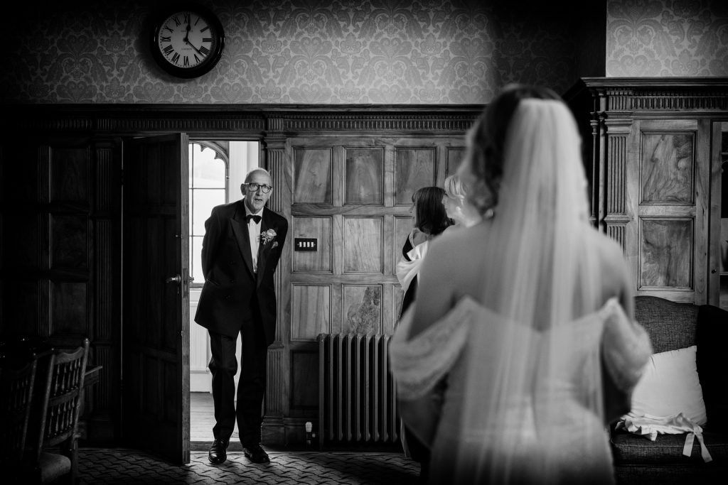 A black-and-white photo shows a man in a suit and bow tie entering a room and smiling at a bride dressed in a wedding gown and veil, who stands with her back to the camera. A clock and wood-paneled walls are visible.