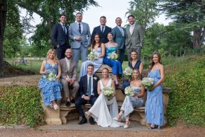 A wedding party poses outdoors on stone steps, with the bride and groom seated in front, bridesmaids in blue dresses, and groomsmen in suits, surrounded by greenery and trees.