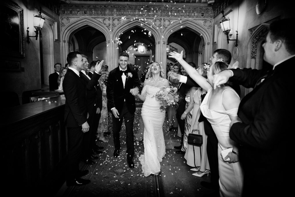 A bride and groom walk down an aisle indoors, smiling as guests on both sides throw flower petals in celebration. The scene is joyful, with elegant clothing and a grand architectural setting. The photo is in black and white.
