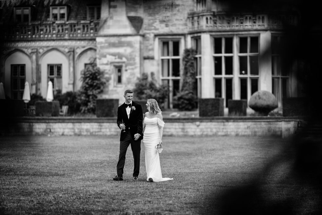 A bride and groom walk hand in hand across a grassy lawn in front of a grand, historic building, both smiling and dressed in formal wedding attire. The photo is in black and white.