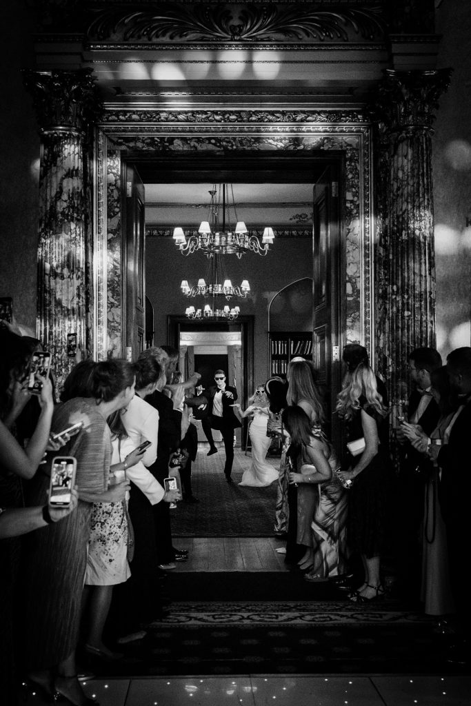 A black-and-white photo of a bride and groom entering an ornate hall, smiling and holding hands, as guests line the hallway, watching and taking photos with their phones. Chandeliers hang above, casting soft light.