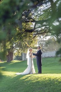 A bride and groom stand facing each other on a sunlit grassy lawn, holding hands beneath a large tree, with soft sunlight filtering through the leaves and a stone wall in the background.