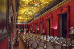 A grand, ornate room with red walls, gold-trimmed ceiling featuring painted murals, and rows of empty elegant chairs arranged for an event; classical statues and large paintings line the room.