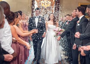 A bride and groom walk down the aisle smiling as guests on both sides throw confetti. The bride wears a white dress and holds a bouquet; the groom wears a black dinner suit. The scene is festive and joyful.