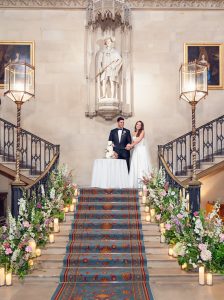 A bride and groom stand at a table on a grand staircase decorated with flower arrangements and lit candles. Ornate lanterns and a statue above them create an elegant setting for their wedding.