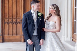 A bride in a strapless white lace gown and a groom in a black suit with a white waistcoat stand together indoors, smiling at each other in front of wooden doors.