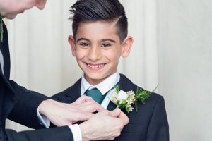 A young boy in a suit and tie smiles as an adult fastens a white buttonhole with greenery to his jacket lapel. The background is plain and light-coloured.