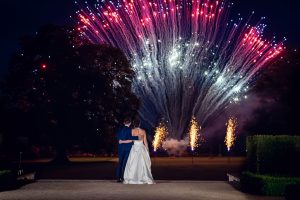 A bride and groom stand arm in arm, facing a vibrant fireworks display lighting up the night sky, with bursts of red, white, and blue above a landscaped garden.
