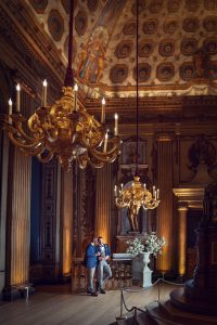 Two people stand closely together in an ornate, high-ceilinged room with golden chandeliers, intricate ceiling art, tall columns, and a large statue, illuminated by warm lighting.