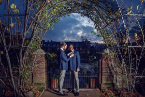 Two men in blue blazers stand under an arched trellis at dusk; one adjusts the other's buttonhole. Behind them, a brick wall and iron gate frame a large building under a dramatic, cloudy evening sky.
