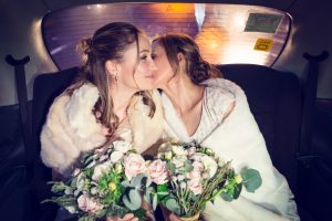 Two brides in white dresses and fur wraps sit closely together in the back of a car, holding bouquets of pink and white flowers, sharing an affectionate moment and smiling.