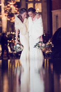 Two brides in white dresses and fur shawls stand close together, smiling and holding bouquets. They are reflected in a shiny surface, with festive lights and flowers in the background.
