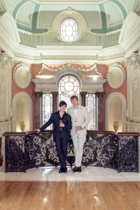 Two people in formal suits stand on an ornate balcony inside an elegant, historic building with decorative columns, statues, and a large arched window behind them. Both are holding drinks and smiling.