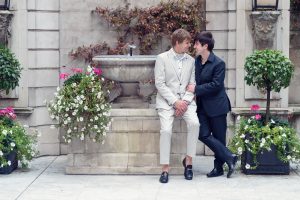 Two men dressed formally, one in a light suit sitting on a stone fountain and the other in a dark suit standing, smile and gaze at each other surrounded by flowering plants in an elegant courtyard.