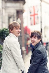 Two men, one in a light suit and the other in a dark jacket, smile and look back whilst holding hands on a city street. A blurred Union Jack flag is visible in the background.