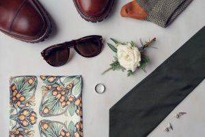 Flat lay of men's accessories including brown shoes, tortoiseshell sunglasses, a floral pocket square, a silver ring, green tie, buttonhole with a white rose, cufflinks, and patterned socks on a light surface.