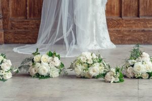 Four white rose bridal bouquets with greenery are arranged on the floor in front of a bride wearing a white lace wedding dress and veil, standing near a wooden wall.