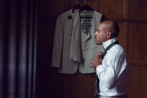 A man in a white shirt stands indoors, adjusting a dark tie. Behind him, a beige suit jacket hangs on a wooden wall. The scene is softly lit, creating a calm and focussed atmosphere.