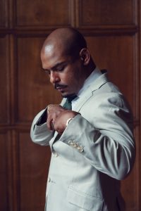 A man in a light grey suit adjusts his jacket, looking down with a thoughtful expression, standing in front of a wooden panelled wall.