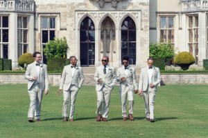 Five men in light suits walk together on a lawn in front of a grand historic building, each holding a drink and smiling, suggesting a celebratory or wedding occasion.