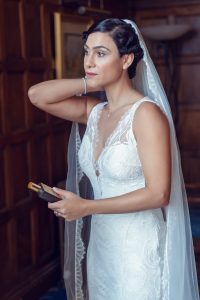 A bride in a white lace wedding dress and veil stands indoors, holding a perfume bottle, with one hand touching her hair and looking thoughtfully to the side.
