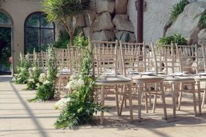 Rows of white chairs are arranged for an indoor wedding ceremony, decorated with green and white floral arrangements. Sunlight streams through large windows, creating a bright and inviting atmosphere.