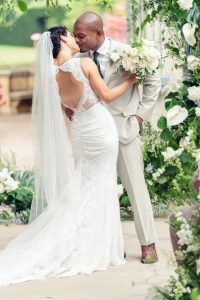 A bride in a lace wedding dress and veil kisses a groom in a light suit. They stand close, surrounded by greenery and white flowers, with the bride holding a bouquet.
