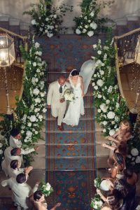 Bride in white gown walks down ornate, flower-lined staircase with her father in a light suit. Wedding guests, some dressed in white, stand on either side, applauding and smiling. Overhead view captures festive, elegant atmosphere.