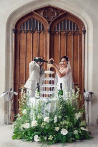 A bride and groom joyfully pour champagne into a tower of glasses in front of large wooden doors, surrounded by white flowers and greenery, celebrating their wedding.
