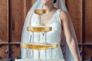 A bride in a white lace dress and veil stands behind a pyramid of filled champagne glasses. The background features wooden panelling. Her face is slightly out of focus and partially out of frame.