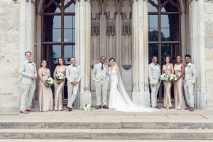 A bride and groom stand with their wedding party in front of a grand stone building with large arched windows. The bridesmaids wear champagne dresses, and the groomsmen wear light grey suits. Everyone is smiling.