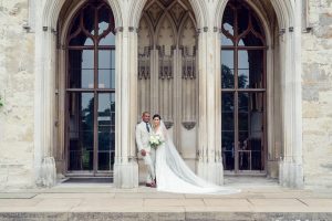 A bride and groom stand smiling together in front of a grand stone building with large arched windows. The bride wears a long white dress and veil, holding a bouquet, whilst the groom wears a light suit.