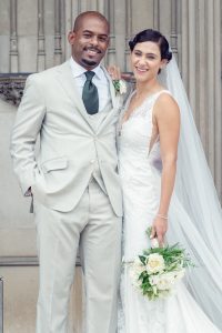 A smiling bride and groom stand together in wedding attire. The groom wears a light grey suit with a dark tie, and the bride wears a white lace dress and holds a bouquet of white flowers, with a long veil draped behind her.