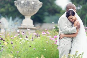 A bride and groom embrace joyfully in a garden filled with wildflowers, with a stone urn and greenery in the background. The bride smiles, wearing a veil, and the groom is dressed in a light-coloured suit.