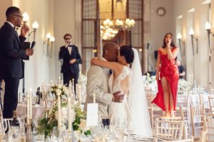 A bride and groom share a kiss during their wedding reception, surrounded by decorated tables. Singers and musicians perform in the background, with a woman in a red dress holding a microphone.