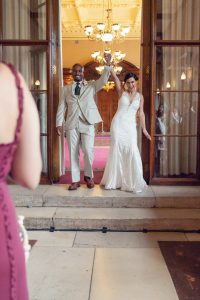 A newlywed couple stands smiling in a doorway, holding hands raised in celebration. The bride wears a white gown, and the groom is in a light suit. Warm, ornate lights glow inside the building behind them.