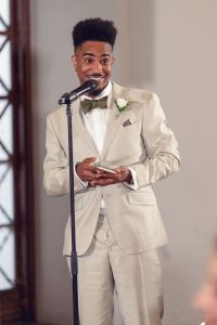 A man in a light beige suit with a green bow tie smiles whilst speaking into a microphone, holding a mobile phone in his hand. He stands indoors near a window, appearing to give a speech at a formal event.