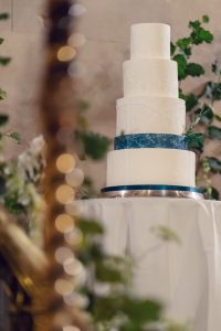 A five-tiered white wedding cake with subtle embossed patterns and a blue ribbon around the bottom tier sits on a round table, surrounded by green foliage and soft, blurred decorative lights in the foreground.