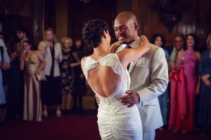 A bride in a white lace dress and a groom in a light suit smile as they dance together, surrounded by guests watching in the background at a formal indoor wedding reception.
