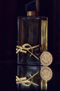 A perfume bottle with gold accents and a gold YSL logo, standing on a reflective surface. The bottle cap is black and gold, and a round wax seal with “YSL” is placed beside the bottle.