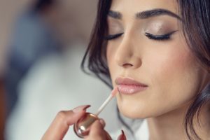 A close-up of a woman with closed eyes having nude lip gloss applied to her lips by another person, highlighting her smooth skin, natural make-up, and subtle winged eyeliner.