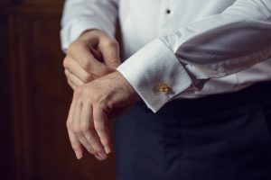 A person in a white dress shirt adjusts a gold cufflink on their sleeve, with both hands visible. The background is softly blurred, highlighting the formal attire.